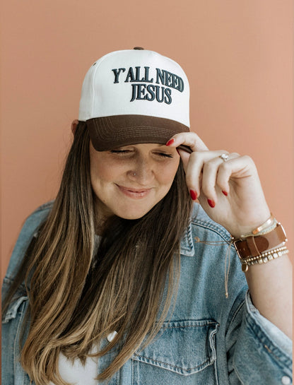 Brown front trucker hat with navy puff embroidery reading “Y’all Need Jesus” — 5-panel snapback style.